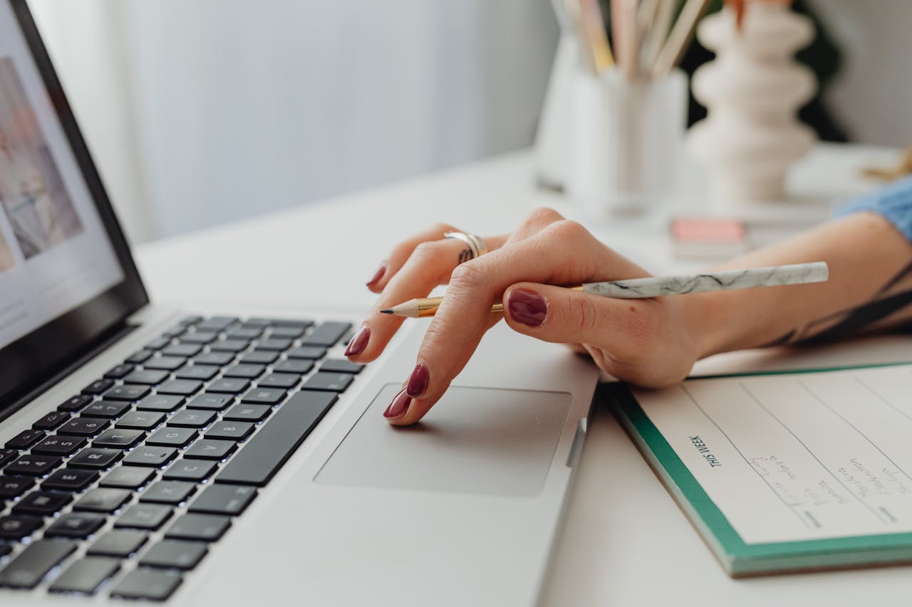 A close-up shot of a womans hand on a laptop touchpad, holding a pencil, beside a notebook.